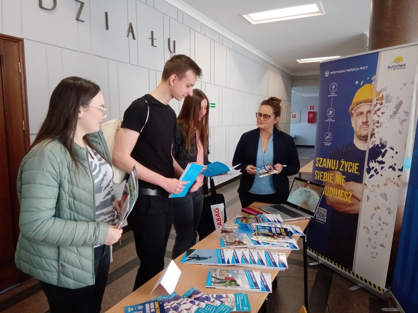 na zdjęciu widać ludzi trzymających ulotki oraz stoisko Panstwowej Inspekcji Pracy, na stole leżą ulotki, obok stołu widać baner z napisem "Szanuj życie, siebie nie odbudujesz"